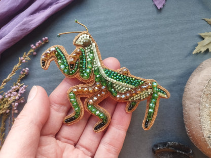 A hand holding a decorative beaded insect, possibly a grasshopper or cricket, against a backdrop of various natural elements such as leaves, flowers, and a stone.