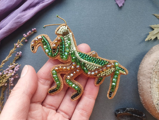 A hand holding a decorative beaded insect, possibly a grasshopper or cricket, against a backdrop of various natural elements such as leaves, flowers, and a stone.