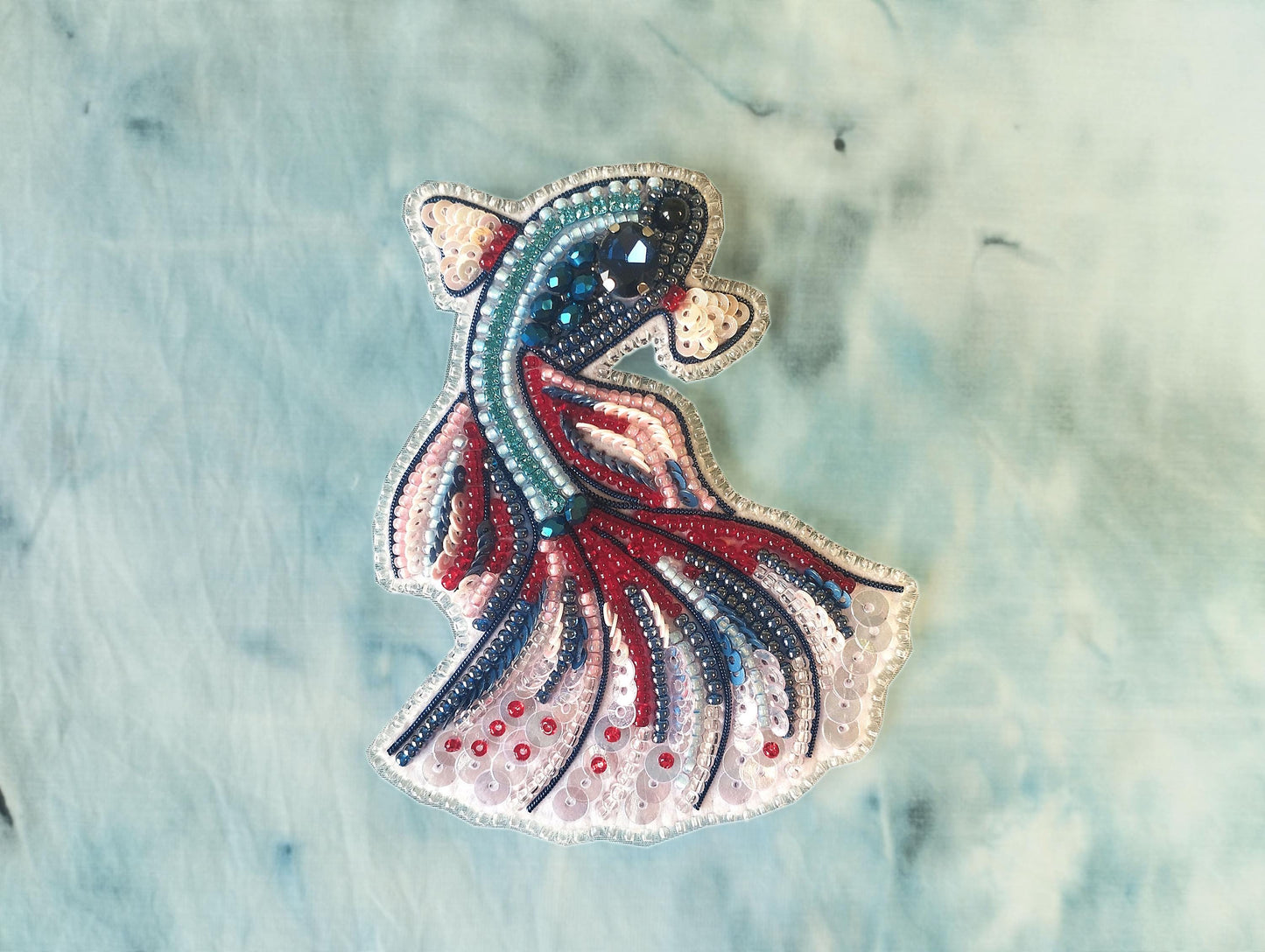 A colorful, intricately designed fish embroidered with various beads and sequins, set against a textured blue background.